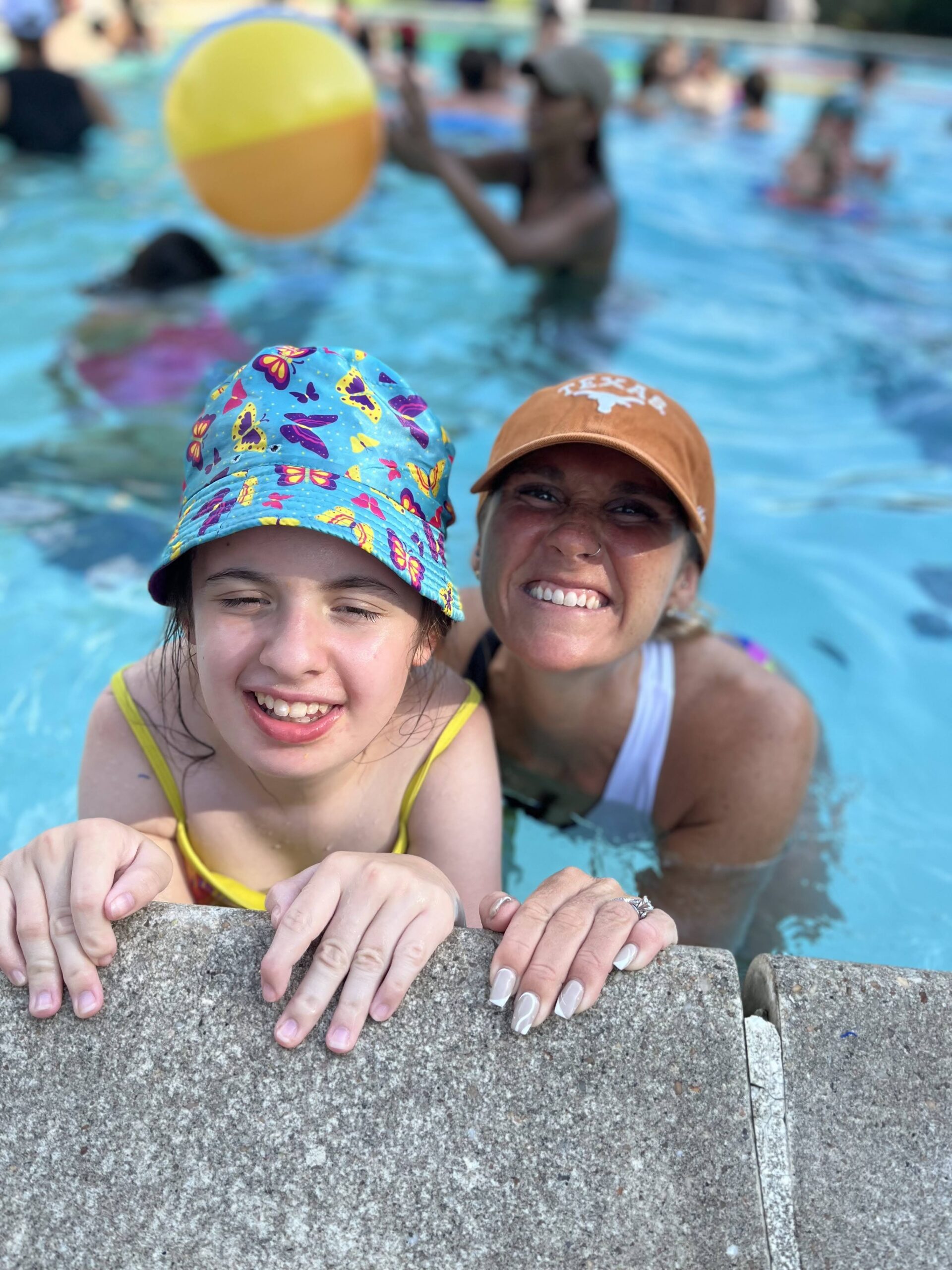 Girl camper in a yellow bathing suit and blue butterfly patterned hat smiling for a picture with a staff member, who is wearing a white tank top and orange baseball cap. 