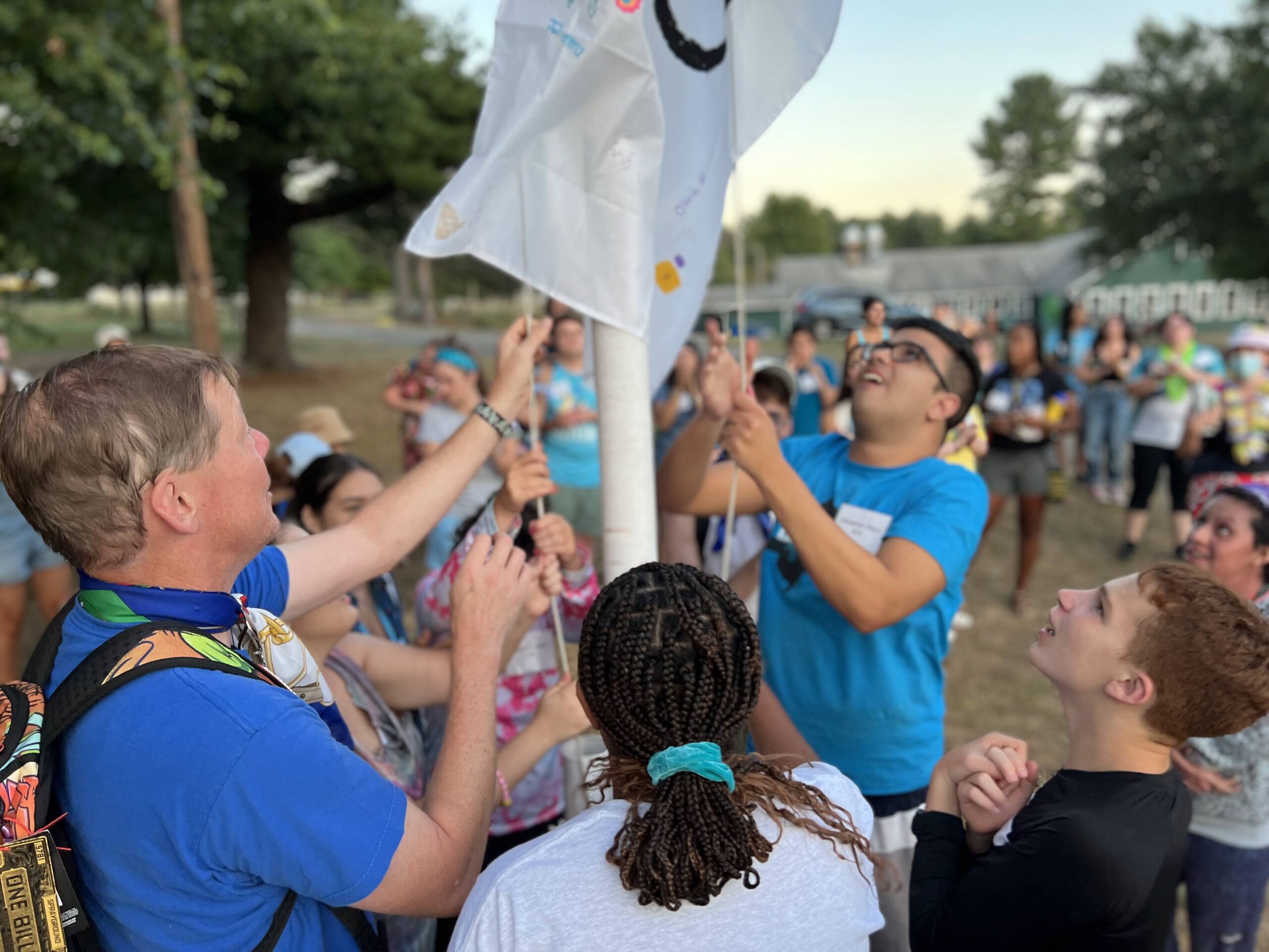 Male camper wearing blue shirt and glasses helping to raise camp flag alongside staff member, also wearing a blue shirt and abstract patterned backpack. 