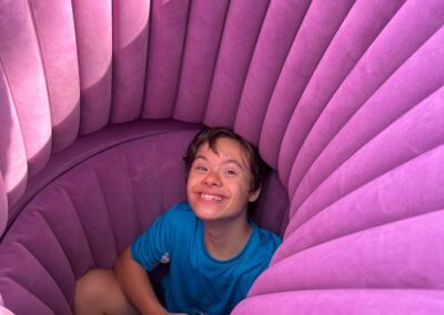 A male camper wearing a blue shirt sitting in a large pink inflatable chair and smiling for the camera.
