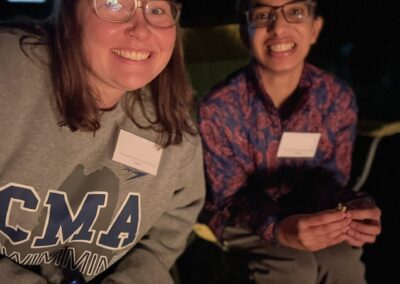 A female staff member wearing a gray sweatshirt and glasses sits next to a female camper wearing a blue and red jacket and black pants. Both are smiling for the camera and sitting beside a campfire, out of frame, at night time.