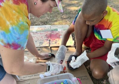 A female staff member wearing a multicolored tie dye shirt and baseball cap kneeling on the ground and helping a male camper wearing a multicolored tie dye shirt and red shorts to make an art project.