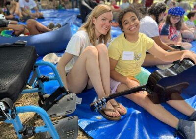 A female staff member wearing a white shirt, jean shorts, and brown flip flops smiling for the camera and sitting on a blue tarp next to a female camper wearing a yellow shirt, pink shorts also smiling for the camera while holding a black acoustic guitar.