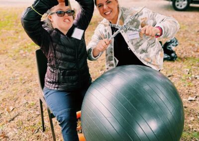 A female camper wearing a black jacket, jeans, and sunglasses is sitting next to a female staff member wearing a black shirt and camo jacket. Both are smiling for the camera and making music by banging on a black yoga ball with drumsticks.